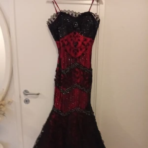 Röd och svart långklänning med spetsdetaljer - Säljer en unik långklänning i rött och svart med smala axelband och spetsdetaljer upptill. Klänningen har figurnära passform, glittriga inslag och dekorativa mönster. Perfekt för bal eller fest! Snygg och dramatisk look.