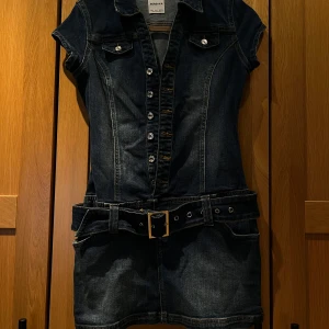 Jeansklänning med bälte från Bershka - Cool jeansklänning från Bershka med korta ärmar, knappar framtill och tillhörande bälte i midjan. Klänningen har klassisk skjortkrage och fickdetaljer på bröstet. Perfekt för dig som gillar denim och vill ha en tuff look.
