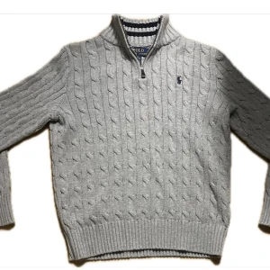 Ljusgrå kabelstickad tröja från Polo Ralph Lauren - Säljer en ljusgrå kabelstickad tröja från Polo Ralph Lauren med halv dragkedja och broderad logga på bröstet. Tröjan har lång ärm, ribbade muddar och klassisk passform. Perfekt för lager-på-lager-stil.