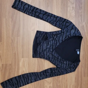 Svart glittrig långärmad topp - Säljer en svart långärmad topp från med glittrigt zebramönster. Toppen har en djup v-ringning och är figurnära i modellen. Perfekt för att sticka ut med lite extra glitter.