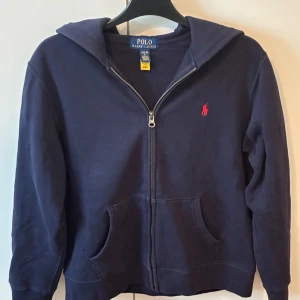 Mörkblå hoodie från Polo Ralph Lauren - Mörkblå hoodie från Polo Ralph Lauren med dragkedja och huva. Klassisk design med röd broderad logga på bröstet och två fickor framtill. Perfekt för en avslappnad stil.