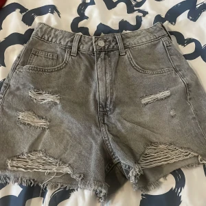 Gråa slitna jeansshorts - Grå jeansshorts med flera slitningar och fransig kant nertill. Klassisk femficksmodell med knapp och dragkedja framtill. Perfekta för en avslappnad stil.