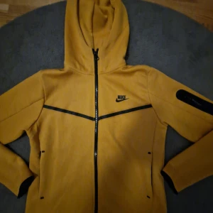 GUL Nike tech fleece storlek: 164 - Säljer en gul hoodiejacka från Nike med svarta detaljer och dragkedja framtill. Jackan har huva, två fickor med dragkedja och en extra ficka på ärmen. Perfekt för träning eller vardag.