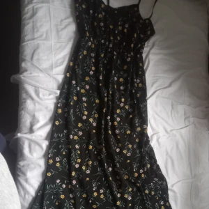 Svart blommig långklänning med smala axelband - Säljer en svart långklänning med smala axelband och blommigt mönster i gult, rosa och grönt. Klänningen har en lätt utställd passform och är perfekt för varma dagar.