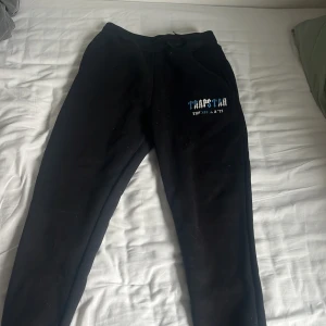 Svarta mjukisbyxor från Trapstar - Svarta mjukisbyxor från Trapstar med tryck i blått och vitt på ena benet där det står 'Trapstar It’s a secret'. Byxorna har en avslappnad passform och resår i midjan.