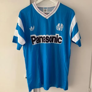 Blå fotbollströja Olympique Marseille från Adidas med Panasonic-tryck - Säljer en klassisk blå fotbollströja från Adidas med Panasonic som sponsortryck på bröstet. Tröjan har vita detaljer på ärmarna och klubbmärke på vänster bröst. V-ringad krage med vita och blå ränder. Perfekt för samlare eller fans! Känns som M i storlek.