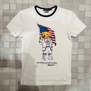 Vit t-shirt med Polo Bear från Ralph Lauren - Vit t-shirt från Ralph Lauren med klassisk Polo Bear-tryck på framsidan där björnen håller en amerikansk flagga och bär vit kostym med en marinblå ribbad krage vilket  ger en snygg look nu till sommaren. Perfekt för dig som gillar en stilren look. Tröjan är nästan aldrig använd och nypris är cirka 1500kr och skick är 10/10.