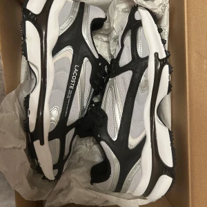 Svarta och vita sneakers från Lacoste - Säljer ett par snygga Lacoste sneakers i svart och vitt med silvriga detaljer. Skorna har snörning, rund tå och en sportig design med tydlig Lacoste-logga på sidan. Perfekta för dig som gillar stilrena och bekväma sneakers.