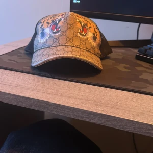 Gucci keps med tigertryck och GG-mönster - Säljer en beige keps från Gucci med klassiskt GG-mönster och detaljerade tigertryck framtill. Kepsen har svart mesh på baksidan och justerbart band med gröna och röda ränder. Perfekt för dig som vill sticka ut med en lyxig accessoar.