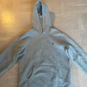 Grå hoodie från Polo Ralph Lauren - Säljer en klassisk grå hoodie från Polo Ralph Lauren med broderad logga på bröstet. Tröjan har huva och en stor magficka framtill. Perfekt för en avslappnad och stilren look. Den är XL i barnstorlek men den passar S!