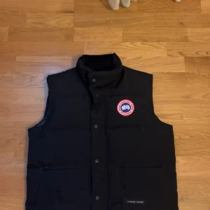 Canada goose väst - Säljer en svart dunväst från Canada Goose med klassisk logotyp på bröstet och hög krage. Västen har knappar framtill och två stora fickor. Perfekt för lager-på-lager och håller dig varm under kyliga dagar.