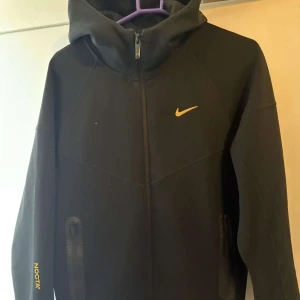 Svart hoodie  Nike NOCTA - Svart hoodie från Nike NOCTA med gul logga på bröstet och NOCTA-tryck på ärmen. 