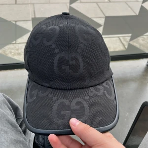 Svart keps från Gucci med GG-mönster - Säljer en svart keps från Gucci med diskret GG-mönster över hela kepsen. Justerbar rem baktill i läderimitation och böjd skärm. Klassisk och stilren design, perfekt för dig som gillar exklusiva accessoarer.