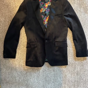 Svart sammetskavaj med färgglatt innerfoder - Snygg svart kavaj i sammet med ett färgglatt, mönstrat innerfoder. Klassisk passform med två fickor framtill och en bröstficka. Perfekt för festliga tillfällen när du vill sticka ut lite extra.