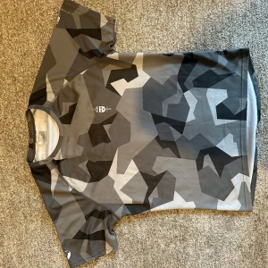 Grå kamouflagemönstrad t-shirt från Trimtex Försvarsmakten - Säljer en grå och svart kamouflagemönstrad t-shirt från Försvarsmakten. T-shirten har rund halsringning och tryckt logga på bröstet samt text på ryggen. Perfekt för dig som gillar militärstil.