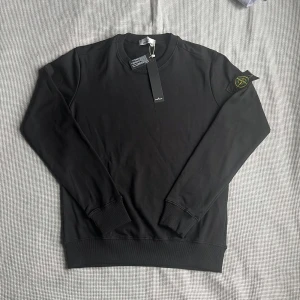 Svart sweatshirt från Stone Island - Säljer en svart sweatshirt från Stone Island med klassisk rund hals och ribbade muddar. Tröjan har den ikoniska Stone Island-loggan på vänster ärm. Perfekt för en stilren och enkel look.