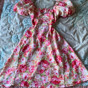 Blommig långklänning med puffärmar - Superfin långklänning med stora puffärmar och färgglatt blommönster i rosa, rött och grönt. Klänningen har v-ringning och öppen rygg med rosettdetalj. Perfekt för sommarens alla tillfällen!