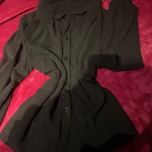 Svart blus med knappar - Säljer en svart blus med knappar framtill och lång ärm. Blusen har diskret struktur i tyget och klassisk krage. Perfekt att styla med kostym byxor eller kjol för en snygg look.