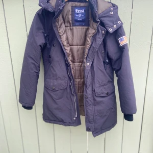 Svart parkajacka från Svea - Säljer en mörkblå parkajacka från Svea med huva och patch med amerikansk flagga på ärmen. Jackan har dragkedja, knappar och flera fickor framtill. Fodrad insida i beige och ribbade muddar vid ärmslut.vindtät. Har en till nästan likadana 