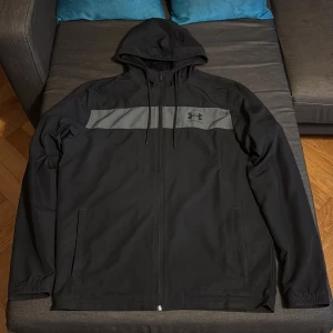 Svart vindjacka med huva från Under Armour - Säljer en svart vindjacka från Under Armour i storlek M. Jackan har huva, dragkedja framtill och en grå panel med logga över bröstet. Två sidofickor och meshfoder på insidan. Perfekt för träning eller vardagsbruk.
