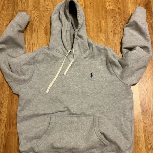 Grå hoodie från Ralph Lauren - Klassisk grå hoodie från Ralph Lauren med broderad blå logga på bröstet. Tröjan har huva med vita snören och en stor magficka framtill. Perfekt för en avslappnad och stilren look.