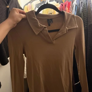 Beige ribbad långärmad topp med krage från Vero Moda - Säljer en beige ribbad långärmad topp från Vero Moda med klassisk krage och v-ringning. Perfekt för en stilren och trendig look. Snygg att matcha med både jeans och kjol!