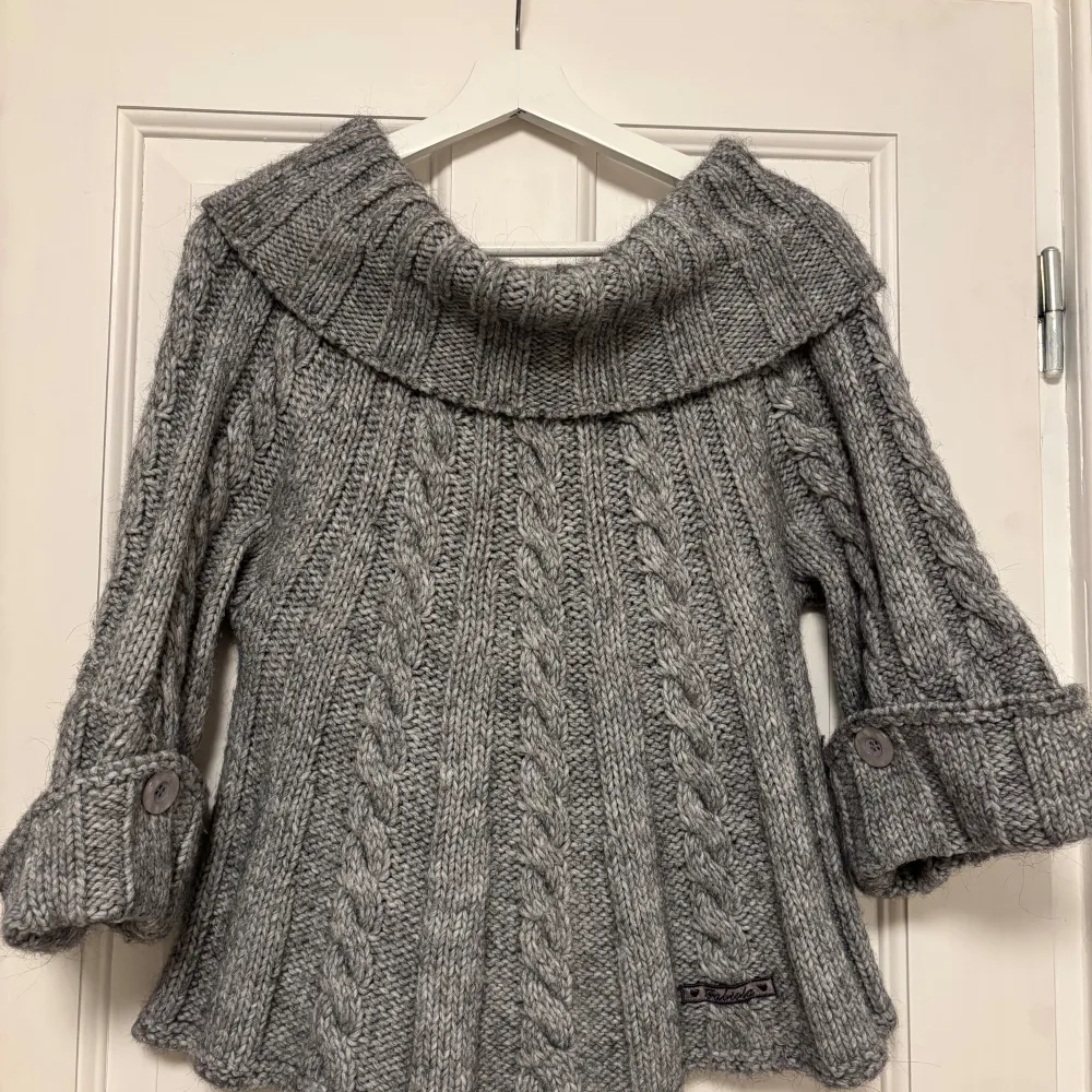 Superfin grå stickad tröja med bred offshoulderkrage. Tröjan har trekvartsärmar med stora uppvik och dekorativa knappar. Sitter löst så kan passa många! Sitter bara åt vid axlarna. 15%ull 15%mohair. Neuletakit & Villapaidat.