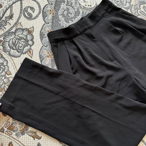 Svarta kostymbyxor från H&M - Klassiska svarta kostymbyxor från H&M med hög midja och raka ben. Byxorna har diskreta veck framtill och en stilren look som passar till många olika outfits.