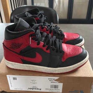 Nike Air Jordan 1 Mid Svart/Röd - Säljer ett par Nike Air Jordan 1 Mid i klassisk svart och röd färgkombination. Skorna har svart bas med röda detaljer, vit mellansula och röd yttersula. Ikonisk Air Jordan-logga på sidan och svart snörning. Perfekta för dig som gillar streetwear och sneakers.