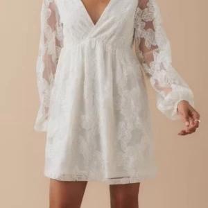 Vit klänning - Superfin vit kortklänning med v-ringad omlott och långa ärmar i transparent spets. Klänningen har ett romantiskt blommigt spetsmönster och lätt utsvängd kjol. Perfekt för studenten 