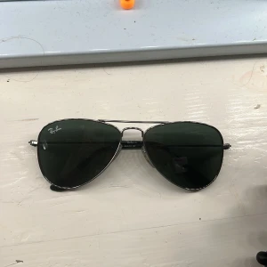 Ray-Ban pilotbågar solglasögon - Klassiska Ray-Ban solglasögon i pilotmodell med tunna silvriga metallbågar och mörkgröna glas. Snygg och tidlös design som passar till många stilar. Ray-Ban-loggan syns på ena glaset. De är väldigt små och rekommenderas för barn eller om man har ett mindre huvud/ansikte.
