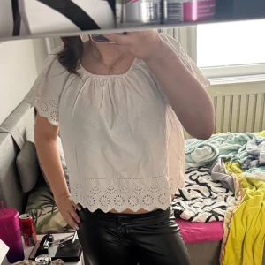 Vit blus med broderade detaljer - Säljer en söt vit blus med korta ärmar och broderade spetsdetaljer längs nederkanten och ärmsluten. Blusen har en lös passform och rundad halsringning med rynk. Perfekt för vår och sommar!