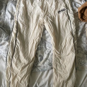 Byxor - Säljer ett par beige träningsbyxor från FSBN med svart 'chaotic system'-tryck på ena benet. Byxorna har svarta detaljer längs sidorna och resår i midjan. Perfekta för träning eller chill. Lätta och luftiga med meshfoder. Använd en gång.