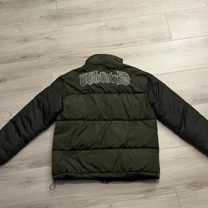 Mörkgrön pufferjacka från BoohooMAN - Säljer en mörkgrön pufferjacka från BoohooMAN med broderad text på ryggen. Jackan har dragkedja framtill, hög krage och två sidofickor. Perfekt för kyliga dagar och enkel att matcha med olika outfits. Jackan är använd BARA vid 1 tillfälle, alltså helt ny