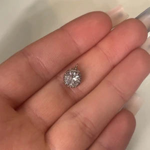 Silverfärgat hängsmycke med glittrig sten - Säljer ett runt hängsmycke i silverfärg med en stor glittrig sten i mitten, omgiven av mindre stenar runt kanten. Perfekt att hänga på en kedja för en elegant look.