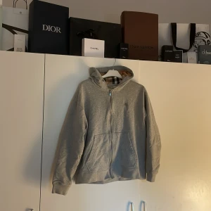 Grå hoodie med dragkedja från Burberry - Säljer en grå hoodie från Burberry med dragkedja och klassiskt rutigt foder i huvan. Tröjan har broderad logga på bröstet och två fickor framtill. Perfekt för en stilren och avslappnad look.