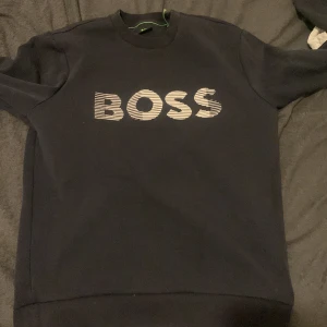 Svart sweatshirt från BOSS - Säljer en svart sweatshirt från BOSS med tryckt logga i vitt på bröstet. Tröjan har rund halsringning och långa ärmar. Perfekt för en stilren och avslappnad look.