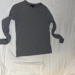 Grå långärmad tröja från Tommy Hilfiger - Säljer en stilren grå långärmad tröja från Tommy Hilfiger. Tröjan har en diskret logga på bröstet och är tillverkad i ett mjukt material. Perfekt för en clean och klassisk look. Lite stöter i storlek så om ni har s så kan ni nog ha den.