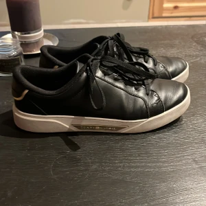 Svarta sneakers från Tommy Hilfiger - Säljer ett par svarta sneakers från Tommy Hilfiger med vita sulor och gulddetalj vid hälen. Skorna har snörning och är tillverkade i skinnliknande material. Perfekta för en stilren och enkel look.