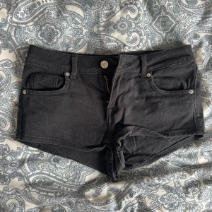 Svarta jeansshorts - Svarta lågmidjade jeansshorts med fickor och knappgylf. Perfekta nu inför sommaren  och enkla att matcha med olika toppar. De är i storlek 34 och i stretchiga material.