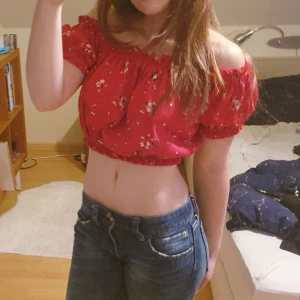 Röd blommig croppad blus - Säljer en söt röd croppad blus med blommigt mönster och volangdetaljer. Toppen har korta puffärmar och resår vid axlar, ärmslut och nederkant. Perfekt för varma dagar och ger en romantisk och söt känsla till outfiten.