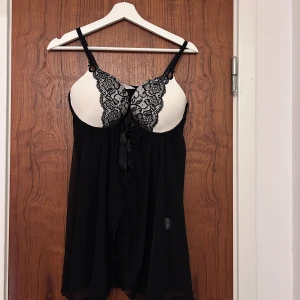 Svart baby doll nattlinne  från Hunkemöller - Säljer en svart babydoll-klänning från Hunkemöller med vita kupor dekorerade med svart spets och smala axelband. Klänningen har en lätt och luftig känsla, perfekt för att känna sig extra fin hemma.