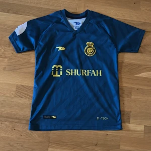 Blå Al Nassr fotbollströja Ronaldo 7 - Snygg blå Al Nassr fotbollströja med guldiga detaljer och tryck. Framsidan har klubbmärke och sponsorlogga i gult, baksidan har namnet Ronaldo och nummer 7. Kortärmad modell, perfekt för fotbollsfans. Säljer pga att den inte kommer till användning 