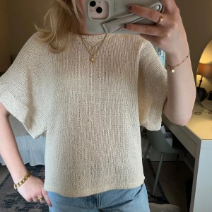 Beige stickad topp från Zara - Säljer en beige stickad topp från Zara som är lite har lite guldiga glitter i. Tröjan har korta ärmar och en avslappnad passform, perfekt till jeans eller kjol. 
