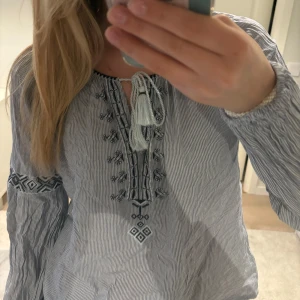 Blåvit blus  - Säljer en blåvit blus från Abercrombie & Fitch med tunna ränder och fina broderade detaljer framtill. Blusen har långa ärmar, knytning vid halsen och dekorativa tofsar. Perfekt för vår och sommar!