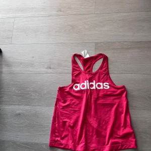 Rosa träningslinne från Adidas - Säljer ett rosa träningslinne från Adidas med vit logga framtill. Linnet har brottarrygg och är gjort i ett lätt och luftigt material, perfekt för träning eller gym. Snygg och sportig design!