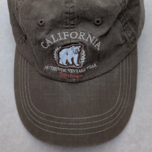 Brun keps med broderad björn och text - Säljer en brun keps med broderad björn och texten 'California Authentic Vintage Wear' framtill. Kepsen har böjd skärm och justerbar rem baktill. Perfekt för en avslappnad stil. Kepsen har aldrig använts 