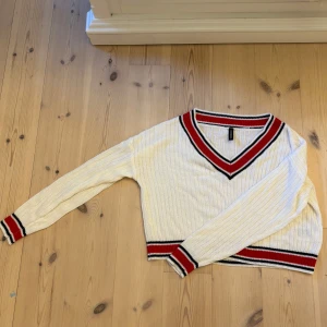 Vit ribbad stickad tröja - Vit stickad tröja från H&M med ribbad struktur och sportiga ränder i rött och svart vid v-ringad hals, ärmslut och midja. Långärmad och croppad modell.