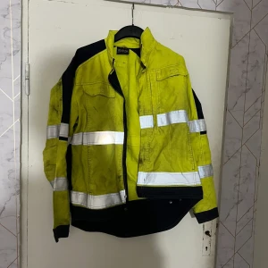 Gul Tranemo varseljacka Strukton Rail L - Varseljacka från Tranemo Workwear i storlek L, gul med svarta detaljer och reflexband runt bröst, armar och rygg. Strukton Rail-logga på ryggen. Jackan har dragkedja framtill, hög krage och flera fickor. Materialmix av bomull och syntet, perfekt för synlighet.