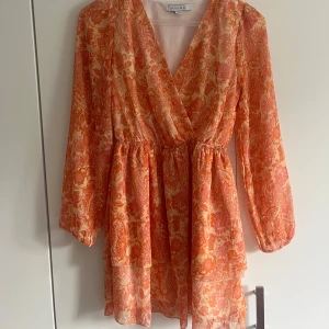 Mönstrad klänning från Amisu - Superfin kort omlottklänning från Amisu i orange och rosa med paisleymönster och subtila glittriga detaljer. Klänningen har långa ärmar, markerad midja och volangkant nertill. Perfekt för dig som gillar färg och mönster!